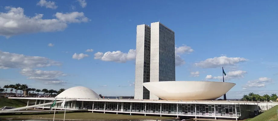 Fotografia do Congresso Nacional do Brasil em um dia ensolarado, com céu azul e nuvens esparsas. A imagem mostra as duas torres gêmeas ao centro, com as cúpulas do Senado Federal (à esquerda) e da Câmara dos Deputados (à direita), símbolos arquitetônicos de Brasília.