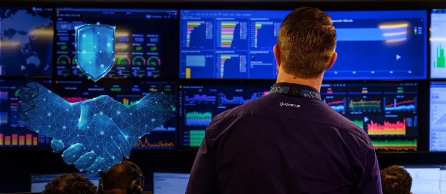 Profissional da Kryptus de costas observando o painel de monitoramento do SOC (Centro de Operações de Segurança) da empresa em Brasília, com múltiplas telas exibindo gráficos, indicadores e dados de segurança cibernética. À esquerda, elementos gráficos em azul representam um aperto de mãos e um escudo digital, simbolizando confiança e proteção. Ambiente moderno e tecnológico.