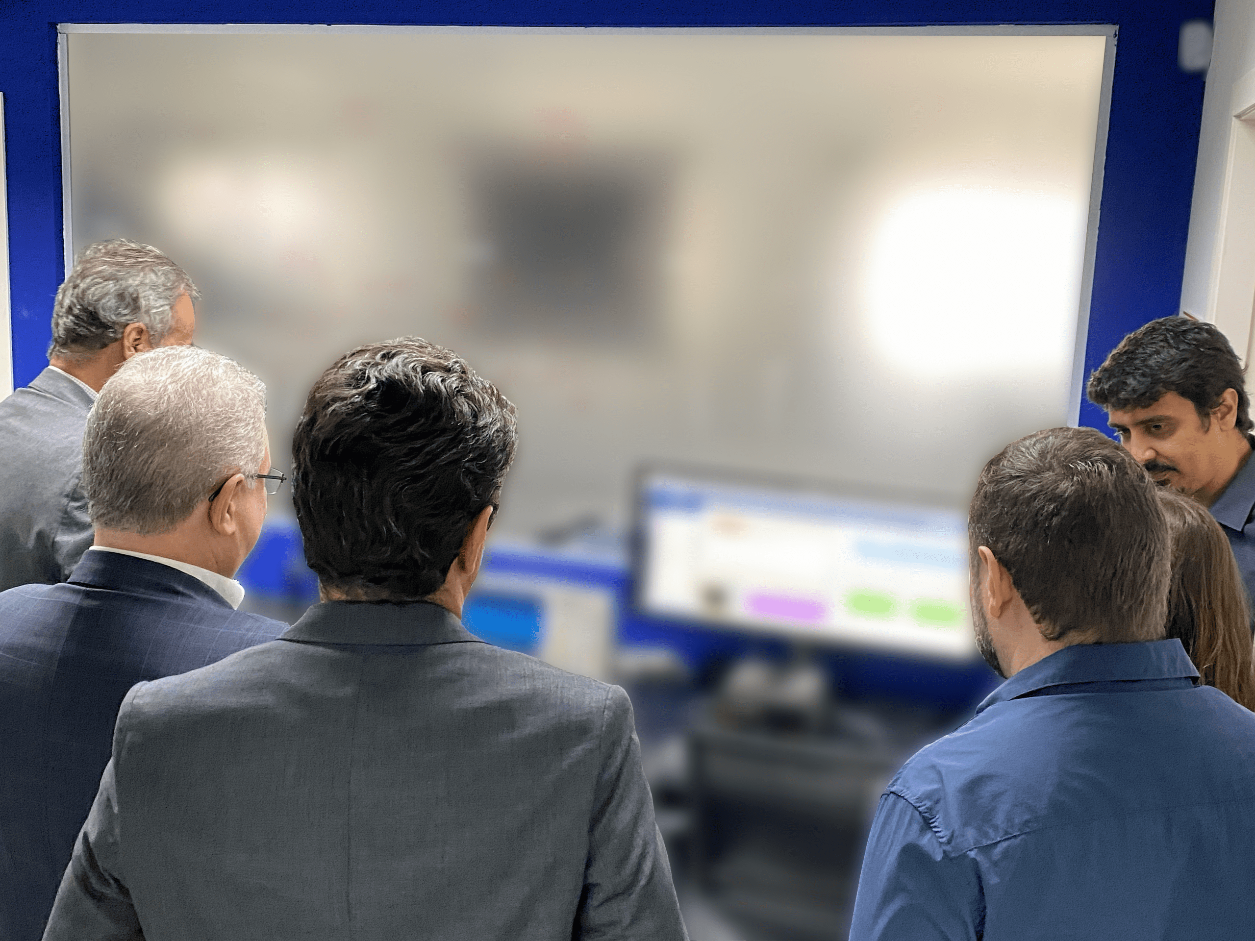 Um grupo de seis pessoas, a maioria homens vestidos com trajes sociais (blazers e camisas), estão em pé e de costas para o observador em uma sala de laboratório com paredes azuis. Eles estão olhando para um grande monitor retangular branco ou tela de projeção desfocada à sua frente, que parece ser um quadro interativo ou tela de visualização de dados. Um monitor de computador, parcialmente visível e desfocado, também está à direita. A cena é uma visita ao LabSCA (Laboratório de Side-channel analysis da Kryptus).