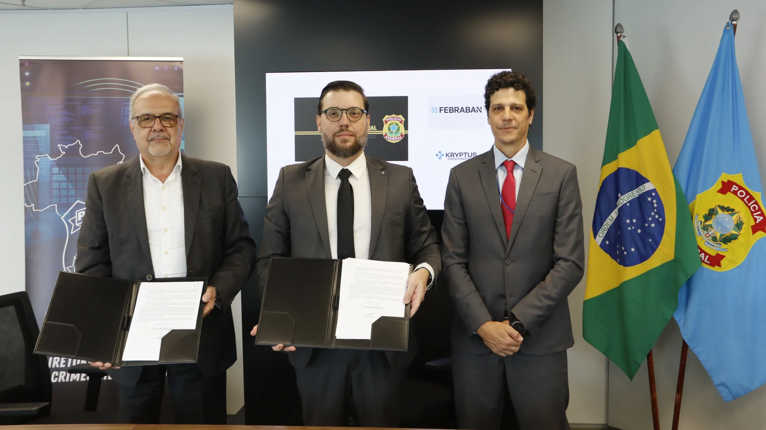 Três homens em trajes formais posam em um escritório em frente a uma tela com os logotipos da Polícia Federal, Febraban e Kryptus. À esquerda, o Diretor-adjunto de Serviços e Segurança da Febraban, Walter Tadeu Pinto de Faria, e ao centro, o CEO da Kryptus, Dr. Roberto Gallo, seguram pastas pretas abertas exibindo as páginas assinadas do contrato do projeto Tentáculos. À direita, está o Diretor de Combate a Crimes Cibernéticos, Otavio Margonari Russo. Ao fundo, à direita, estão posicionadas as bandeiras do Brasil e da Polícia Federal.